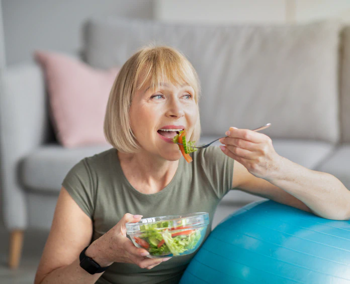 A smiling woman eating a fresh salad near an exercise ball, highlighting the role of cleanse and detox supplements in a healthy lifestyle.