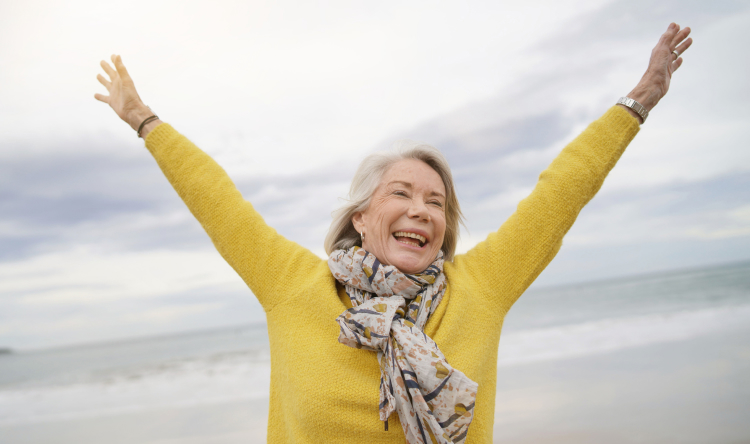 A joyful elderly woman raising her arms at the beach, representing the benefits of supplements for energy and vitality.