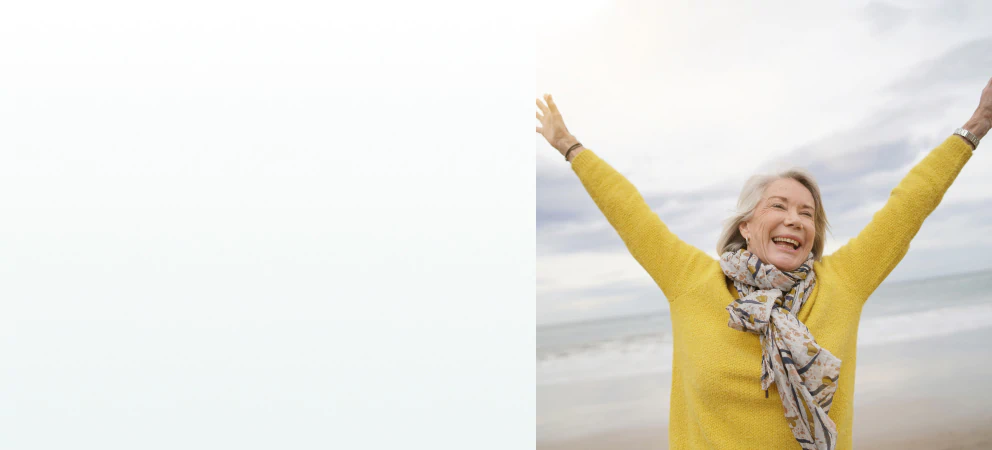 A joyful elderly woman raising her arms at the beach, representing the benefits of supplements for energy and vitality.