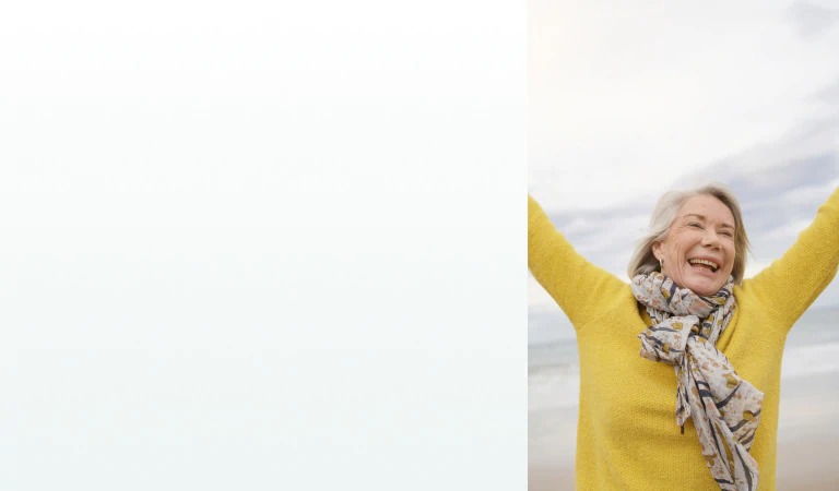 A joyful elderly woman raising her arms at the beach, representing the benefits of supplements for energy and vitality.