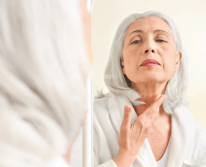 A mature woman applying skincare to her neck while looking in the mirror, reflecting the role of skin and beauty supplements.
