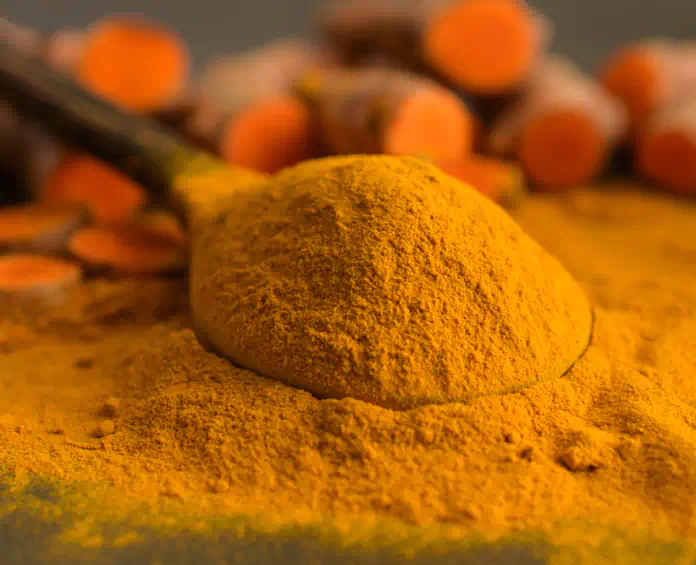A spoonful of bright yellow turmeric powder with fresh turmeric roots in the background, a key ingredient in toenail fungus supplements.