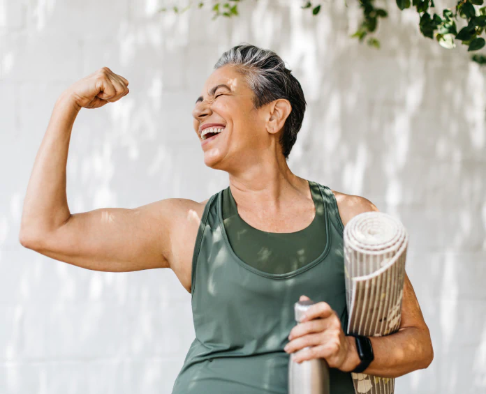 A fit, smiling woman flexing her arm while holding a yoga mat, showing strength and well-being with the help of women's health supplements.