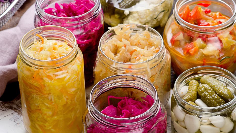 A top-down image showing multiple glass jars filled with different pickled vegetables, including pink sauerkraut, yellow sauerkraut, pickled peppers.